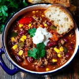 Eine Schüssel mit herzhaftem Chili, garniert mit Sauerrahm und frischen Korianderblättern, neben einer Scheibe geröstetem Brot.