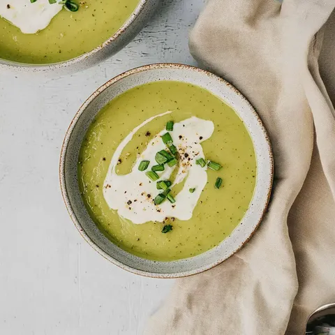 Zwei Schalen mit grüner Suppe, garniert mit Sahne, Schnittlauch und Pfeffer, auf einer hellen Unterlage.