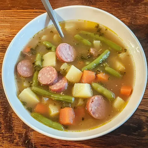 Eine Schüssel mit Gemüsesuppe, die Würstchen, grüne Bohnen, Karotten und Kartoffelstücke enthält.