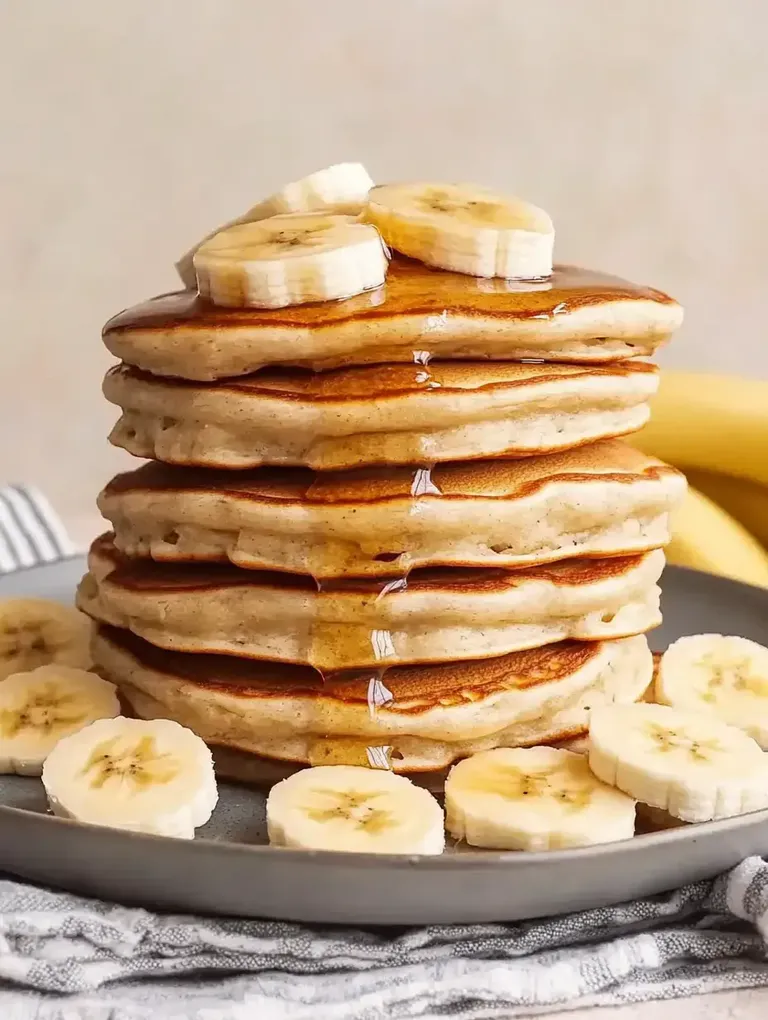 Ein Stapel Pfannkuchen mit Bananenscheiben und Sirup, angerichtet auf einem grauen Teller.